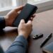 Unas manos sostienen un teléfono móvil, mientras una billetera física para criptomonedas se encuentra al lado.